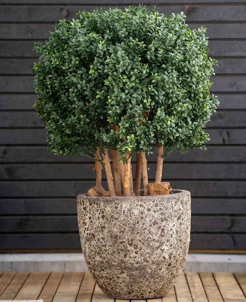 La palla di bosso artificiale in legno stile romano UV 90 cm, una pianta rotonda a foglia verde con steli spessi, si trova in un grande vaso di pietra su legno, contro una parete orizzontale scura con pannelli di legno.