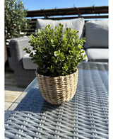 La pianta artificiale Buxus verde in vaso UV 19 cm, con foglie verdi in un vaso a forma di cesto di vimini, poggia su un tavolo da esterno in vetro con mobili da giardino e una ringhiera in legno visibile sotto un cielo azzurro e limpido.