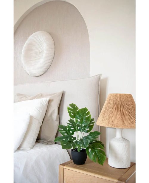 Una camera da letto moderna dai toni neutri ha un letto bianco con cuscini beige e bianchi, un comodino in legno con una lampada in ceramica e un paralume intrecciato, quadri astratti sopra il letto e Art Monstera 40 cm in un vaso decorativo nero che aggiunge verde.