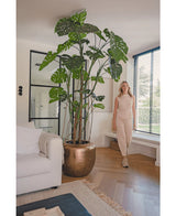 Una donna vestita di beige sta accanto alla pianta artificiale Monstera alta 240 cm in un vaso di bronzo. Il soggiorno moderno ha un divano bianco, grandi finestre con persiane e un pavimento in legno.