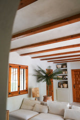 Un accogliente soggiorno con pareti bianche, travi in legno, un divano componibile bianco, la palma artificiale Phoenix De Luxe da 210 cm, finestre e porte in legno, scaffali integrati con cestini e ceramiche e luce naturale che entra dalla finestra.