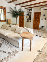 Un accogliente soggiorno con divano bianco, tavolino rotondo in legno, palma artificiale Phoenix De Luxe 210 cm, cestini intrecciati su scaffali, porte in legno, uno specchio rotondo e tappeti dai toni neutri su un pavimento chiaro.