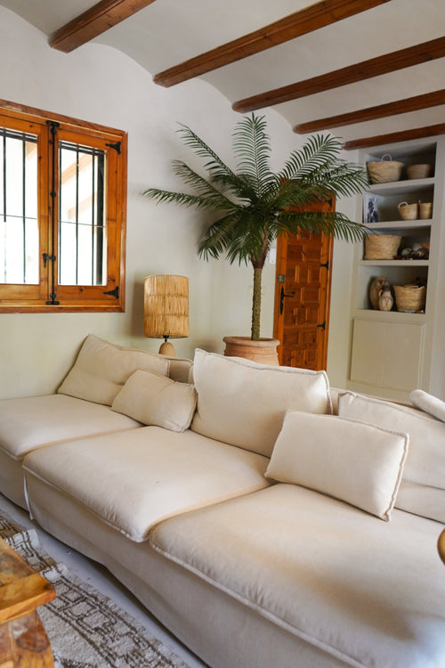Un accogliente soggiorno con un grande divano color crema, la Palma Artificiale Phoenix De Luxe 210 cm nell'angolo, una lampada in legno, scaffali integrati con cestini, travi in legno e una finestra con cornici in legno.