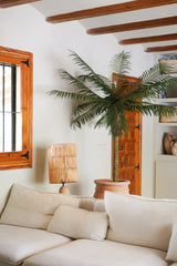 Un accogliente soggiorno con un divano bianco, una lampada da tavolo in rattan, la palma artificiale Phoenix De Luxe da 210 cm in un vaso, travi in legno sul soffitto, una finestra con cornici in legno e una porta in legno sullo sfondo.