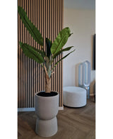 Il Banano Artificiale 150 cm, con le sue foglie verdi lussureggianti, si erge su un pavimento di legno vicino ad una parete nervata con pannelli di legno. Nelle vicinanze c'è un pouf rotondo bianco e un ventilatore senza pale sullo sfondo.