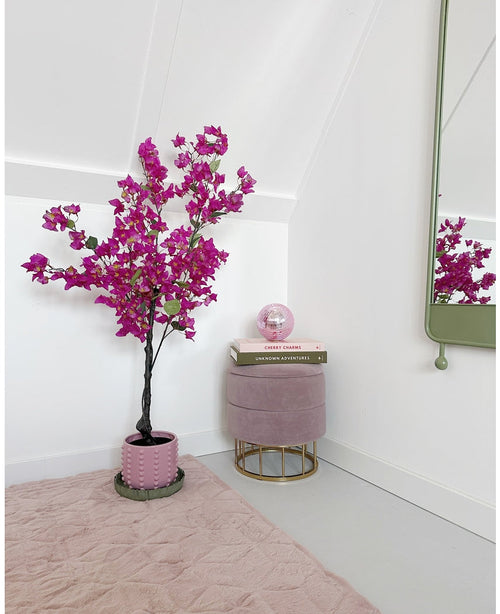 Un angolo moderno e minimale con una pianta di Bougainvillea artificiale viola da 125 cm in vaso, un poggiapiedi rotondo rosa con gambe dorate, libri impilati, una sfera di cristallo rosa, uno specchio a parete e un tappeto rosa tenue su un pavimento chiaro.