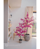 Due bouganville artificiali viola 125 cm con vivaci fiori viola in vasi strutturati illuminano una stanza moderna, accanto a una sedia in legno, una libreria e una grande finestra con tende beige.