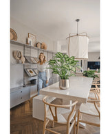 Una donna sorride in una sala da pranzo moderna e luminosa con sedie in legno, un tavolo bianco con la pianta artificiale Dieffenbachia da 75 cm come centrotavola e scaffali aperti decorati con libri e opere d'arte.