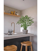 Una cucina moderna con un piano di lavoro in cemento, due sgabelli in legno e una pianta di Dieffenbachia artificiale di 75 cm accanto al rubinetto in ottone, oltre a dispenser di sapone e una mensola in legno con vasi contro una parete grigio chiaro.