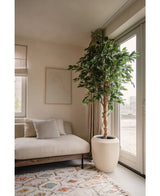 Un soggiorno moderno con un divano beige, un cuscino a righe, quadri astratti, un tappeto a motivi, una finestra a tutta altezza con tende trasparenti che lasciano entrare la luce naturale e una pianta artificiale Ficus Verde 210 cm.