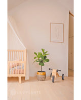 Una cameretta minimale con un letto in legno chiaro, un baldacchino traslucido, una pianta artificiale di ficus tabacco Deluxe da 90 cm, giocattoli in legno con l'etichetta Mia e una stampa incorniciata in stile line art minimalista su una semplice parete bianca.