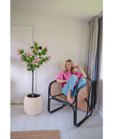 Una donna con un maglione rosa e jeans blu siede a piedi nudi su una sedia in rattan accanto alla Pianta Artificiale Giuda 150 cm, un albero artificiale con fiori rosa, in una stanza moderna e luminosa con pareti e pavimenti bianchi.