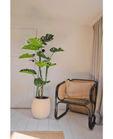 Una pianta artificiale Monstera di 150 cm si trova accanto a una sedia in rattan con un cuscino marrone in una stanza luminosa e minimalista con pareti bianche, pavimenti in legno chiaro e tende beige.