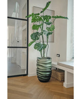 La pianta artificiale Monstera 150 cm, con le sue grandi foglie verdi, si trova in un vaso con motivo ondulato su un pavimento di legno in una stanza moderna e luminosa con una porta a vetri, quadri incorniciati e un cesto di vimini nelle vicinanze.