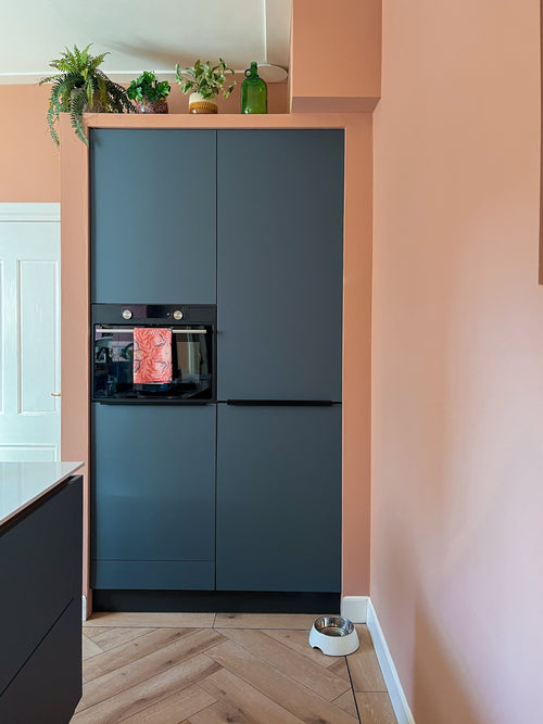 Un angolo cucina moderno con mobili scuri opachi, un forno da incasso e un pavimento in legno chiaro. Sopra i mobili sono posizionati la pianta artificiale Monstera da 18 cm e una bottiglia in vetro verde; una ciotola per il cibo per animali domestici è appoggiata sul muro color pesca.