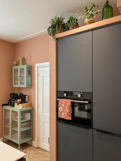 Una cucina moderna con mobili scuri opachi, un forno da incasso e un asciugamano fantasia. Sopra gli armadi ci sono piante in vaso come la pianta artificiale Monstera da 18 cm e bottiglie di vetro, mentre un armadio verde menta nell'angolo contiene le scorte di caffè.