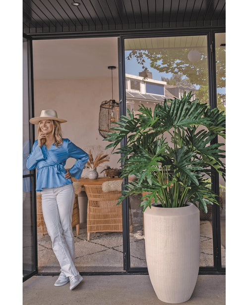Una donna con una camicetta blu, pantaloni bianchi e cappello ride davanti a una porta a vetri accanto alla pianta di filodendro artificiale di 125 cm in un vaso beige. Sedie e decorazioni in vimini sono visibili in tutta la stanza.