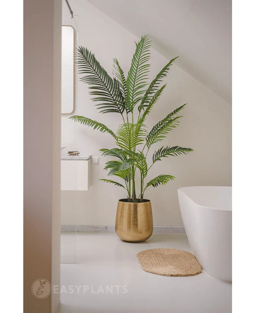 La pianta artificiale Phoenix da 180 cm in un vaso dorato si trova accanto a una vasca da bagno bianca e un lavandino in un bagno moderno e minimalista con pareti e pavimento bianchi. Nelle vicinanze c'è una stuoia rotonda intrecciata sul pavimento.