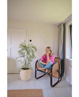 Una donna con una camicia rosa e jeans siede a piedi nudi su una sedia intrecciata, sorridendo e rilassandosi in una stanza luminosa e minimalista con pareti e pavimenti bianchi. Accanto a lei c'è la pianta Artificiale Ruscus 120 cm.