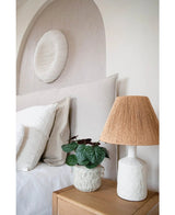 Una camera da letto moderna con una tavolozza di colori neutri ha un letto con lenzuola bianche, un comodino in legno, una lampada strutturata in ceramica con paralume intrecciato, la pianta artificiale Sassifraga 27 cm e una scultura a parete bianca rotonda sopra la testiera.