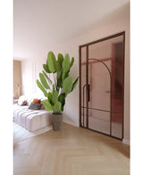 Un soggiorno moderno con divano bianco, cuscini fantasia e la pianta artificiale Strelitzia Real Touch Deluxe 210 cm. Un pavimento in legno chiaro, pareti rosa tenue e una porta in vetro con struttura in metallo bronzo creano un'atmosfera leggera e ariosa.