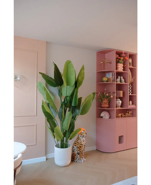 La pianta artificiale Strelitzia Real Touch Deluxe 210 cm si trova accanto a una figura di ghepardo in ceramica con un cappello arancione, accanto a un'alta mensola rosa piena di vasi, fiori e ornamenti in una stanza dai colori pastello.