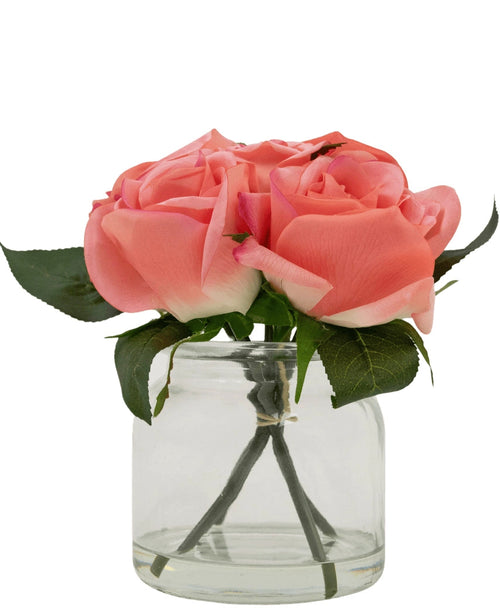 Il bouquet di rose artificiali Real Touch Pink 18 cm contiene quattro rose artificiali rosa con foglie verdi, disposte in un piccolo barattolo di vetro su uno sfondo bianco.