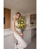 Una donna dai capelli biondi ricci, vestita con un abito color crema, si appoggia a un bancone e tiene in mano il bouquet 'Ramo artificiale di limone 104 cm giallo', decorato con una vegetazione lussureggiante. La cucina moderna è decorata con colori neutri e presenta accenti in legno.