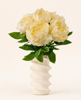 Un vaso bianco ondulato e strutturato contiene il bouquet di fiori artificiali in seta Peonia Bianca: grandi fiori di peonia artificiale color crema con foglie verdi su uno sfondo chiaro e semplice.
