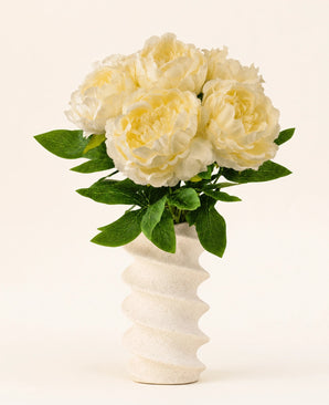 Un vaso bianco ondulato e strutturato contiene il bouquet di fiori artificiali in seta Peonia Bianca: grandi fiori di peonia artificiale color crema con foglie verdi su uno sfondo chiaro e semplice.