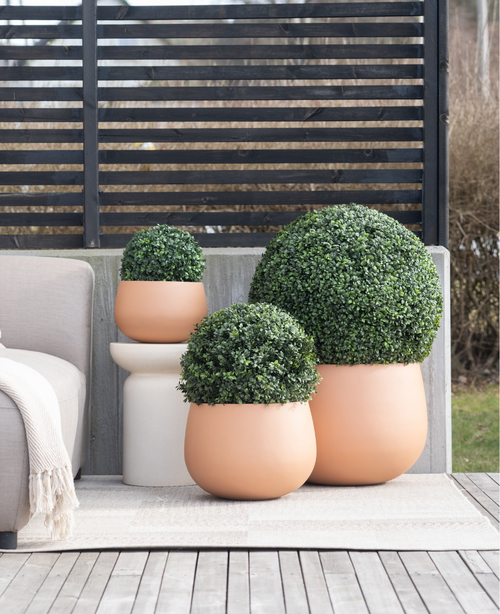 Tre fioriere rotonde in terracotta UV D30 cm in stile romano bosso artificiale con arbusti verdi tagliati si trovano su un patio accanto a un divano da esterno beige e un tavolino bianco, con uno schermo per la privacy con doghe dietro di esso.