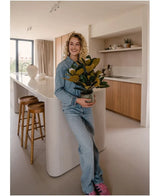 Una persona sorridente vestita di jeans si trova in una cucina moderna con in mano una pianta di magnolia artificiale in un vaso da 46 cm, una pianta di facile manutenzione. La cucina, perfetta per la decorazione d'interni, è dotata di mobili in legno, un'isola bianca e due sgabelli da bar.