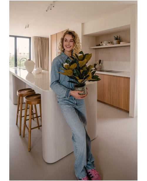 Una persona sorridente vestita di jeans si trova in una cucina moderna con in mano una pianta di magnolia artificiale in un vaso da 46 cm, una pianta di facile manutenzione. La cucina, perfetta per la decorazione d'interni, è dotata di mobili in legno, un'isola bianca e due sgabelli da bar.