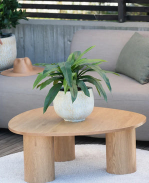 Una pianta artificiale Bird's Deluxe di 60 cm si trova su un tavolino rotondo in legno davanti a un divano beige, decorato con un cuscino verde e un cappello marrone appeso sopra il bracciolo.