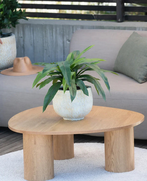 Una pianta artificiale Bird's Deluxe di 60 cm si trova su un tavolino rotondo in legno davanti a un divano beige, decorato con un cuscino verde e un cappello marrone appeso sopra il bracciolo.