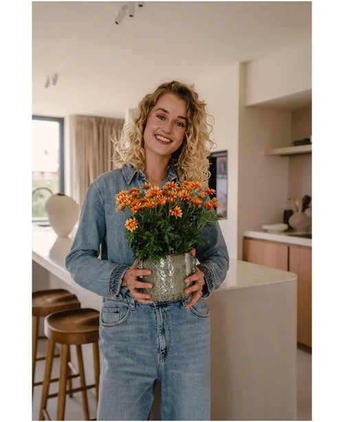 Una donna con i capelli biondi ricci, che indossa abiti in denim, sorride mentre tiene in mano la pianta Artificial Daisy UV 31 cm – un vaso di fiori gialli e arancioni brillanti – in una cucina moderna.