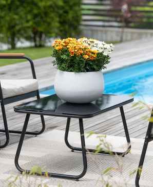 Accanto a una piscina, circondato da sedie da giardino e con il verde sullo sfondo, si trova un tavolo da patio nero con una pianta Artificial Daisy UV da 31 cm - un vaso rotondo bianco con vivaci margherite bianche e arancioni -.
