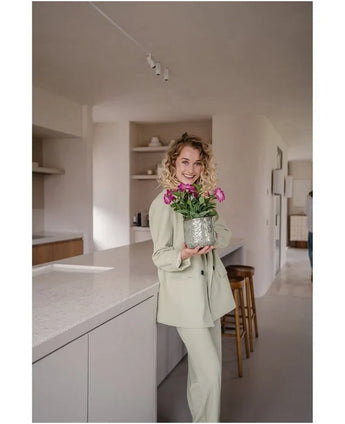 Una donna in abito chiaro sorride mentre tiene in mano un fiore artificiale Petunia UV 25 cm rosa scuro; si trova in una cucina moderna dai toni neutri e dall'arredamento minimalista.