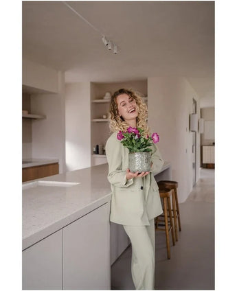 Una donna sorridente con i capelli ricci, che indossa un abito chiaro, sta in una cucina moderna e minimalista con in mano un fiore artificiale Petunia UV 25 cm rosa scuro con fiori rosa vivaci.