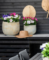 Due grandi vasi in ceramica con un fiore artificiale Petunia UV 25 cm rosa scuro, con fiori rosa vivaci, stanno su un tavolo contro una parete di legno scuro. C'è un cappello di paglia sul tavolo e foglie di palma essiccate sono appese al muro sopra i fiori.