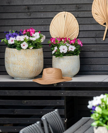 Due grandi vasi in ceramica con un fiore artificiale Petunia UV 25 cm rosa scuro, con fiori rosa vivaci, stanno su un tavolo contro una parete di legno scuro. C'è un cappello di paglia sul tavolo e foglie di palma essiccate sono appese al muro sopra i fiori.