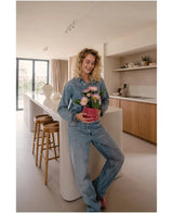 Una donna in jeans è in piedi in una cucina moderna, sorride e tiene tra le mani la pianta di Gerbera artificiale UV rosa da 28 cm: un piccolo vaso di fiori rosa con fiori rosa e bianchi. Sullo sfondo si vedono due sgabelli in legno e una grande finestra.