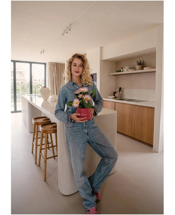 Una persona in abito jeans si trova in una cucina moderna e sorride alla telecamera mentre tiene in mano la pianta artificiale Gerbera UV 28 cm rosa. Intorno all'isola della cucina sono disposti sgabelli in legno e sullo sfondo si possono vedere mensole decorative.