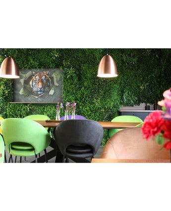 Una moderna sala da pranzo con sedie verdi e color pesca, un tavolo decorato con vasi di fiori viola, lampade a sospensione in rame, una rigogliosa parete vegetale realizzata con il pannello Kunsthaag Crush UV di 100 x 100 cm e un'immagine incorniciata di una tigre sullo sfondo.