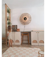 Una giovane ragazza con un vestito leggero sta accanto a uno scaffale su cui stanno la pianta pensile artificiale 'Frosted Fern on ball 76 cm' e altre piante pendenti, in una stanza moderna con armadi beige, uno sgabello alto, una lampada da parete in vimini e un tappeto a quadri sul pavimento.