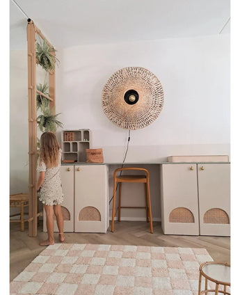 Una giovane ragazza con un vestito leggero sta accanto a uno scaffale su cui stanno la pianta pensile artificiale 'Frosted Fern on ball 76 cm' e altre piante pendenti, in una stanza moderna con armadi beige, uno sgabello alto, una lampada da parete in vimini e un tappeto a quadri sul pavimento.