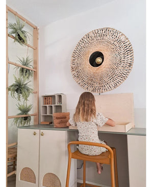 Una bambina con i capelli lunghi fino alle spalle siede a una scrivania in una stanza luminosa, con la schiena rivolta allo spettatore. Dietro di lei ci sono una lampada da parete intrecciata, alcuni armadietti e la pianta artificiale pensile 'Frosted Fern on ball 76 cm', annidata tra le piante su una scala di legno, accanto a piccoli scaffali con oggetti disposti in modo ordinato.
