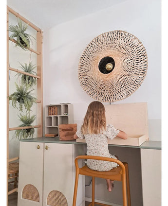 Una bambina con i capelli lunghi fino alle spalle siede a una scrivania in una stanza luminosa, con la schiena rivolta allo spettatore. Dietro di lei ci sono una lampada da parete intrecciata, alcuni armadietti e la pianta artificiale pensile 'Frosted Fern on ball 76 cm', annidata tra le piante su una scala di legno, accanto a piccoli scaffali con oggetti disposti in modo ordinato.