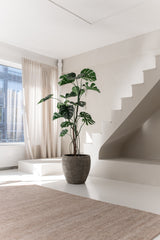 De Kunstplant Monstera 210 cm staat in een pot op een witte vloer naast een moderne trap in een lichte, minimalistische kamer met natuurlijk licht dat door beige gordijnen naar binnen valt.