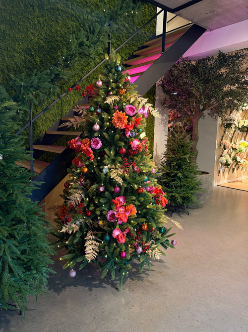 L'Albero di Natale Artificiale "Weideman" PE/PVC - Smart LED si trova in interni vicino ad una scala, decorato con fiori rosa e rossi, foglie dorate e ornamenti rosa, circondato dal verde su un pavimento di cemento lucido per un'elegante decorazione natalizia.